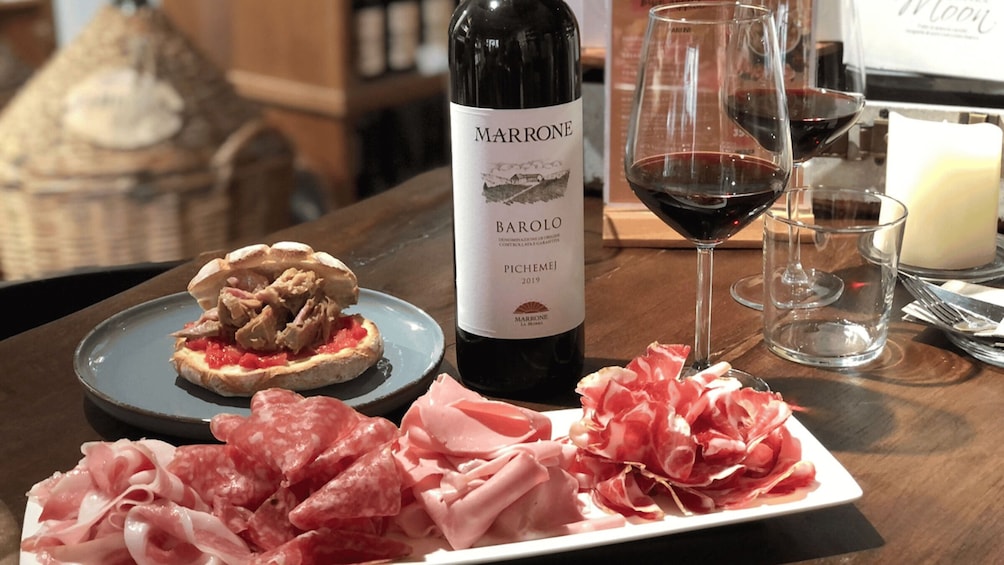 A bottle of Barolo wine, a glass of red wine, and a plate of cured meats on a wooden table. 1