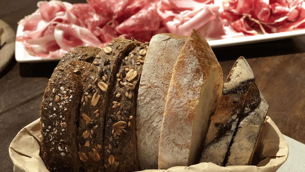 A variety of breads with seeds and grains, and a plate of sliced meats in the background. 3