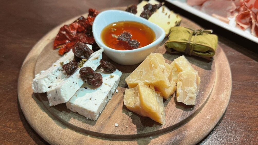 A wooden board with various cheeses, dried fruits, and a bowl of sauce. 3