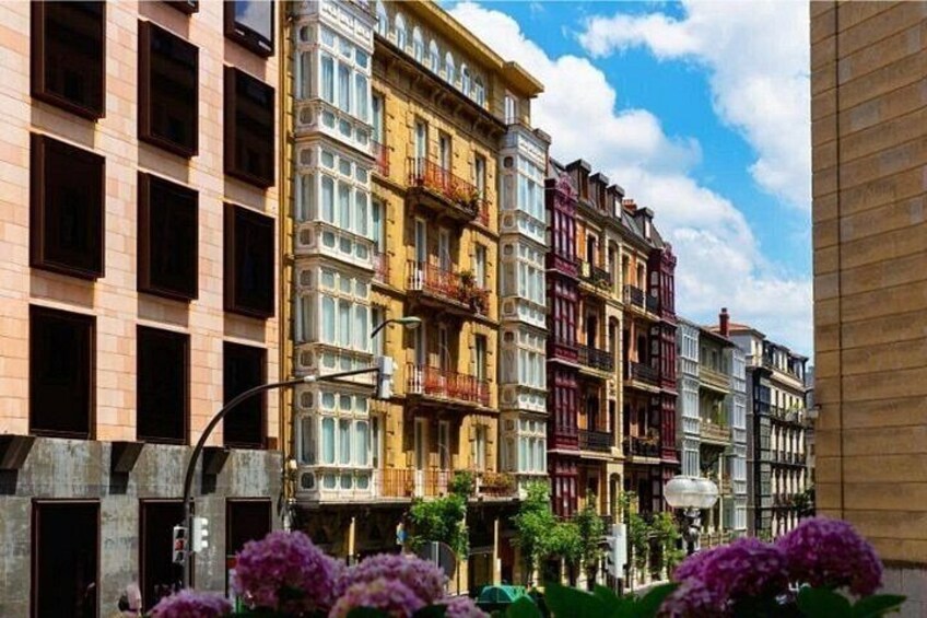 A row of colorful historic buildings with balconies and windows, a clear sky, and purple flowers in the foreground. 2