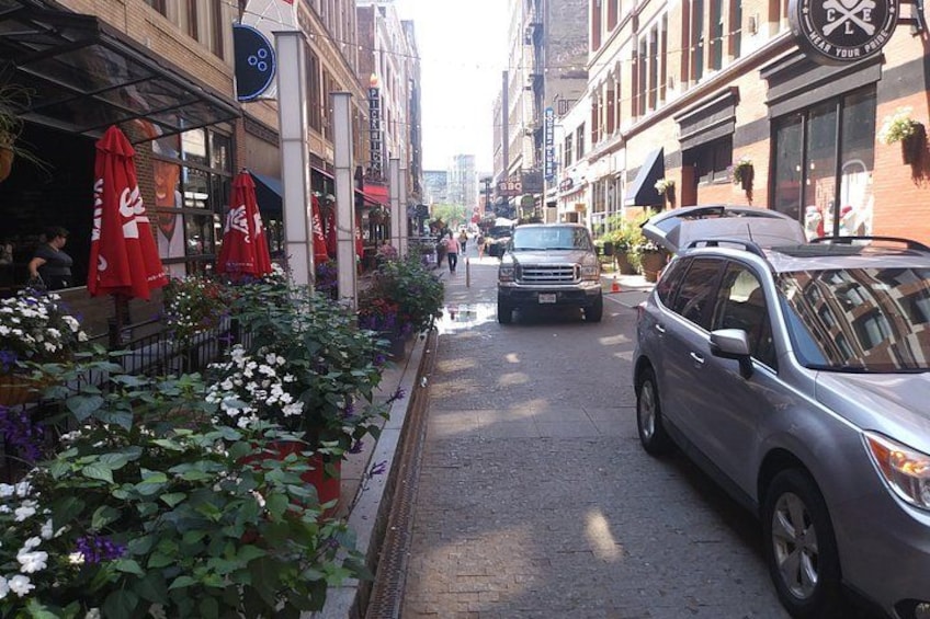 Une rue étroite en milieu urbain, bordée de jardinières et d’ombrelles rouges, avec des voitures garées et un immeuble doté d’une horloge. 1