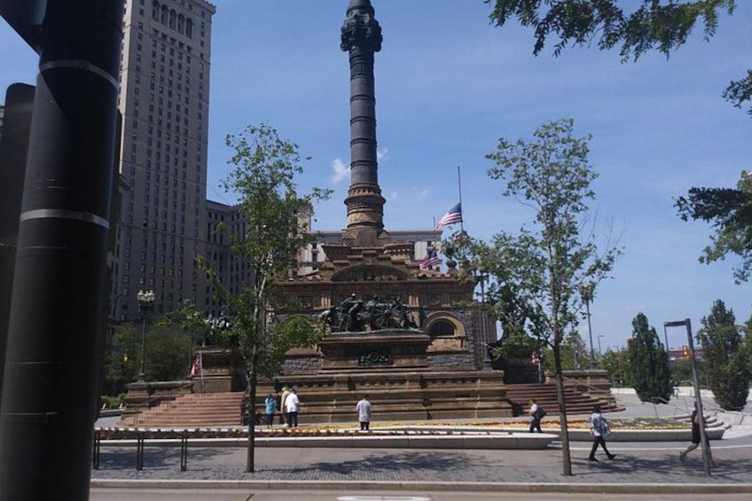Un monument composé d’une haute colonne, entourée d’escaliers et surmontée d’un socle de statue, avec des drapeaux et une vue sur la ville en arrière-plan. 2