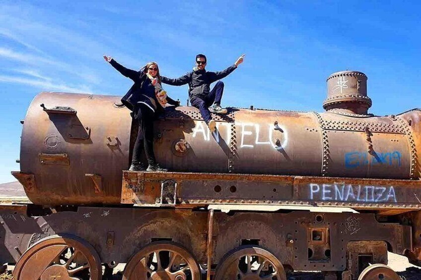 A rusty train engine with graffiti, two people posing on it, and a clear blue sky in the background. 5