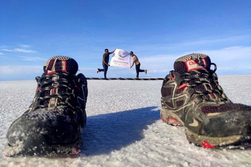 A pair of muddy hiking boots on a snowy surface with a person holding a sign in the background. 1