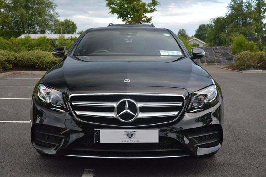 En svart Mercedes-Benz-bil står parkerad på en parkeringsplats med träd och en stenmur i bakgrunden. 1