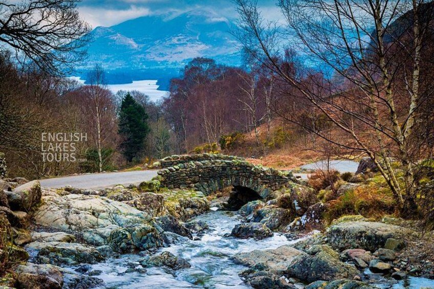 A stone bridge over a flowing stream, surrounded by trees and a distant lake. 3
