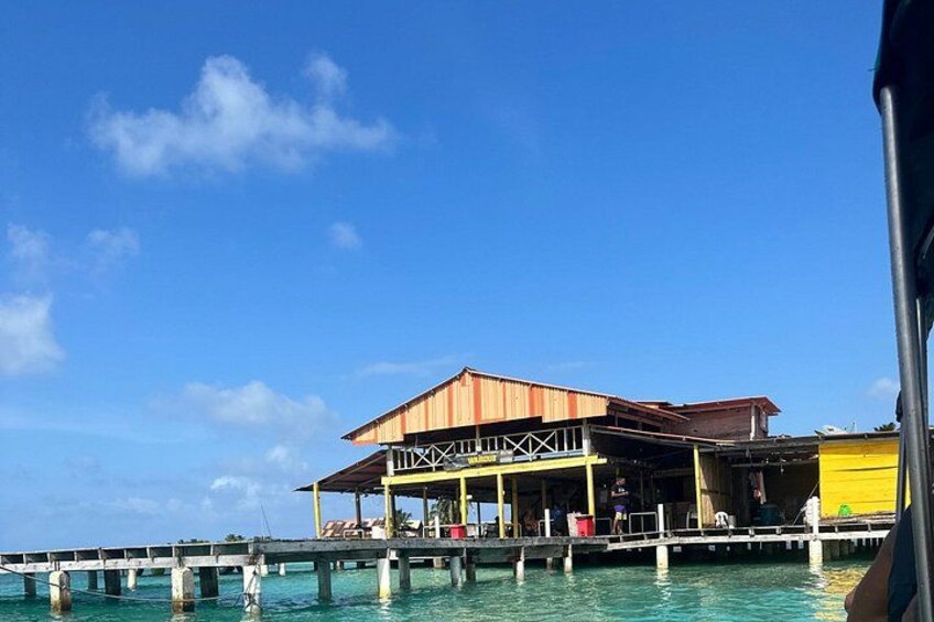 Een houten gebouw met een metalen dak, een steiger die in het turquoise water uitloopt, en een helderblauwe lucht. 4