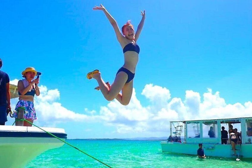 A person jumping off a boat into turquoise water, a boat with people on it, and a clear blue sky. 2