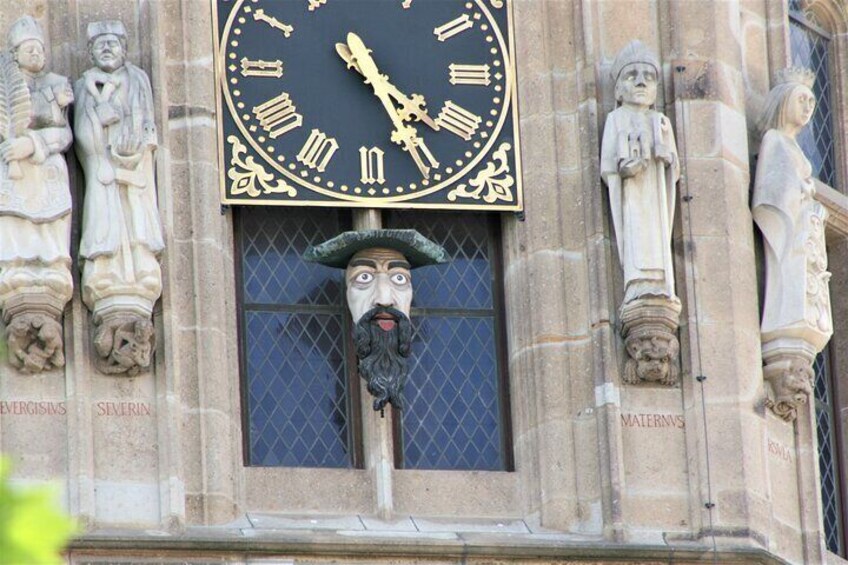 A clock with Roman numerals and a statue of a man with a beard in front of it. 4