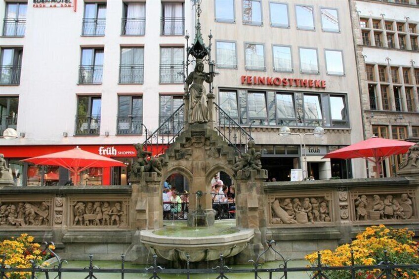 A fountain with sculptures in front of a building with a sign that reads "FEINKOSTHEKE". 2
