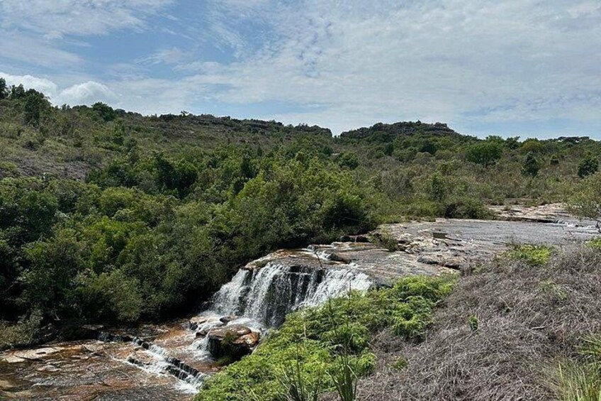 A waterfall cascading over rocks in a forested area with dense greenery and a hilly backdrop. 2
