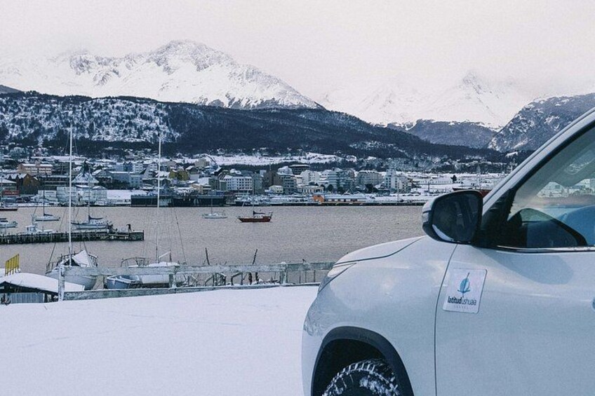 A snowy landscape with a car parked in the foreground, a marina with boats, and snow-covered mountains in the background. 4