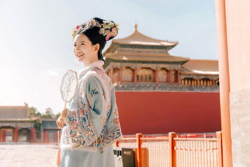 A person in traditional attire holding a fan, standing in front of a historical building with a distinctive roof. 1