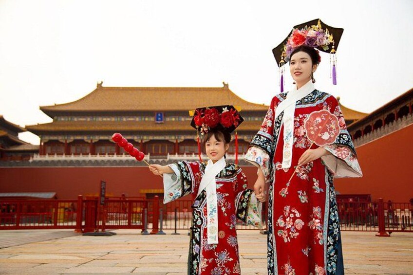 Two individuals in traditional attire, a child and an adult, standing in front of a historic building with a tiled roof and red walls. 2
