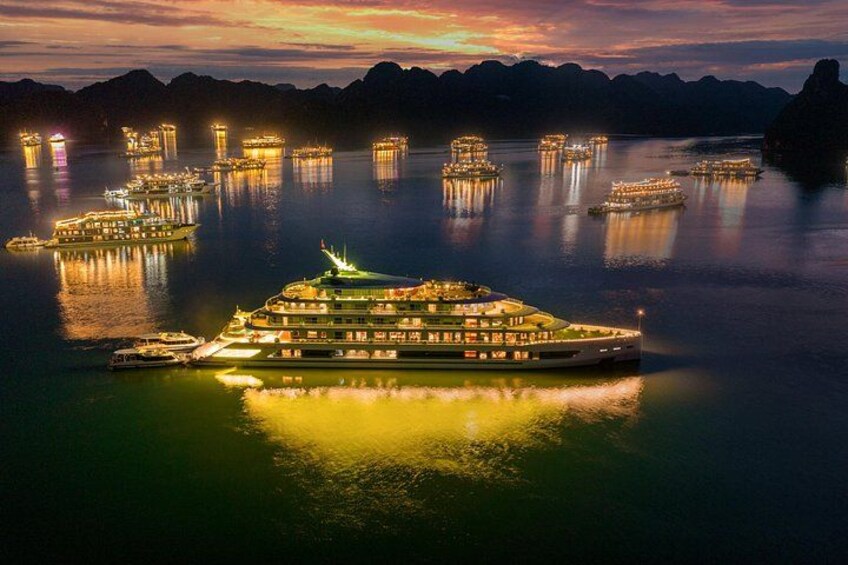 A large illuminated boat with multiple decks docked at a harbor surrounded by smaller boats and structures. 2