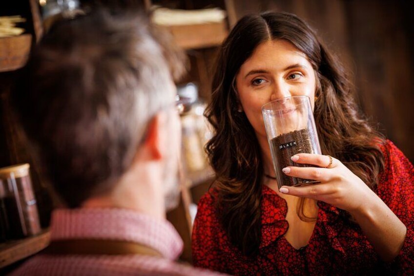 A person holding a glass with a dark liquid, likely coffee or tea, in a setting with wooden shelves and jars. 2
