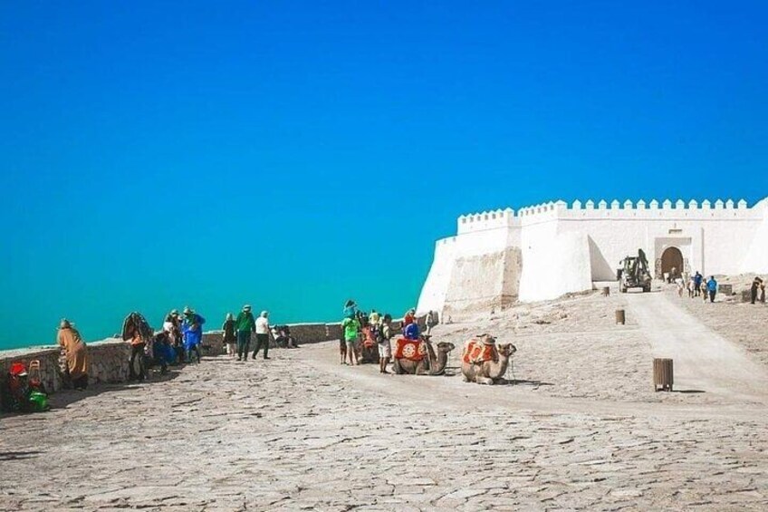 A coastal area with a white fort-like structure, a sandy path, and people walking. 2