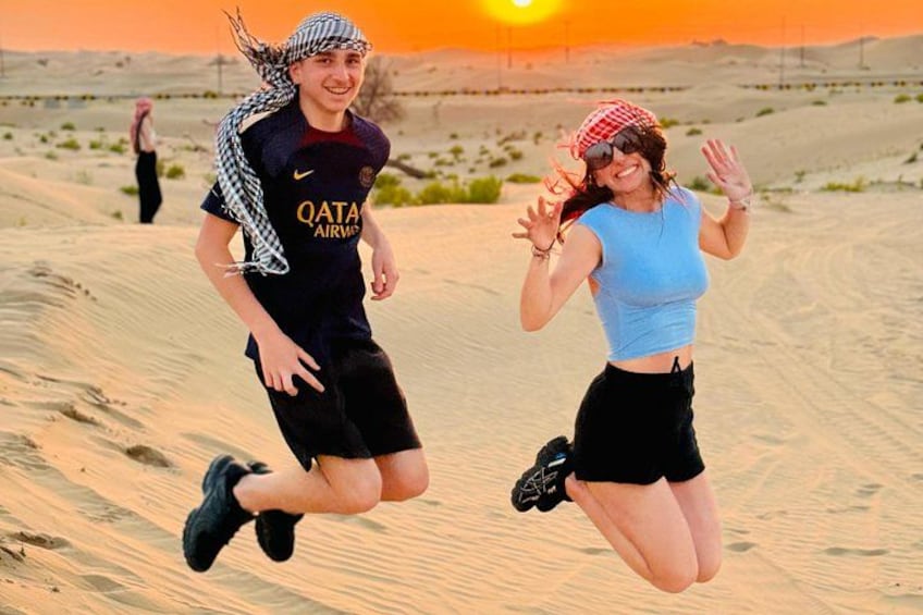 Due persone che saltano in un deserto con dune di sabbia e un tramonto sullo sfondo. 5