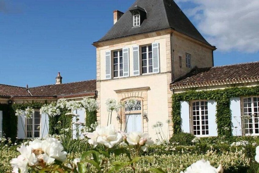 A two-story house with a gabled roof, a front door with a sign, and a garden with white flowers. 2