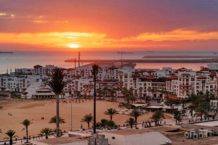 A coastal cityscape at sunset with buildings, palm trees, and a beach. 2