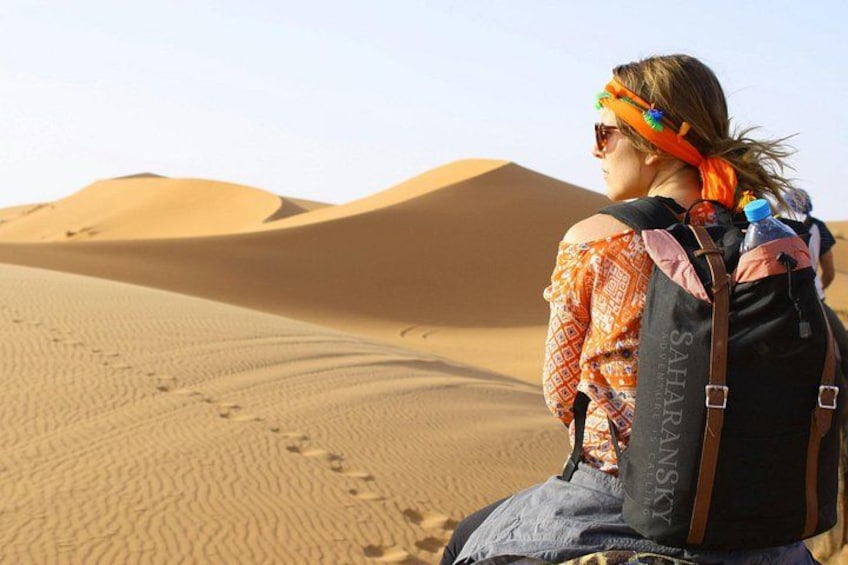 A person sitting on a sand dune with a backpack and a patterned shirt. 2