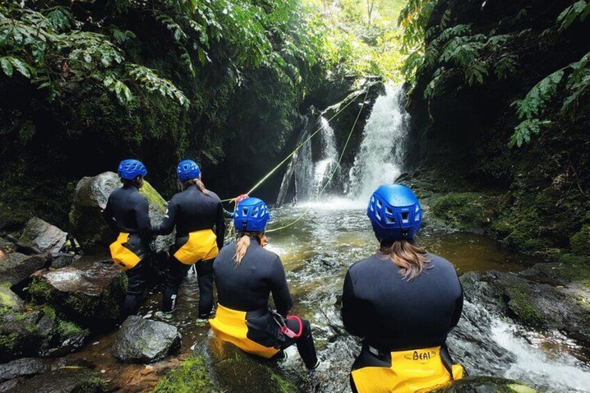 Een groep mensen met helmen en neopreen pakken staat bij een waterval in een bebost gebied. 1