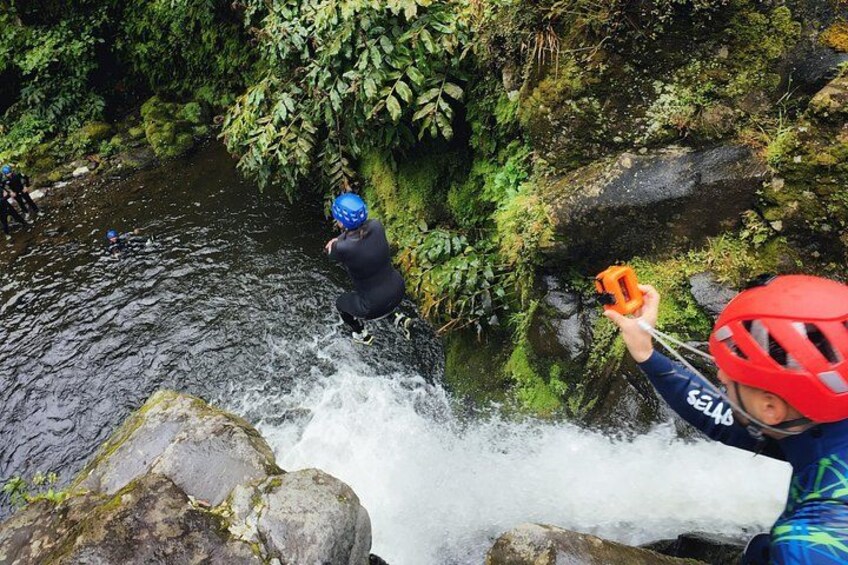 A person in a blue wetsuit and red helmet near a waterfall, another person in a black wetsuit and blue helmet, and a third person in a blue wetsuit and orange helmet. 3