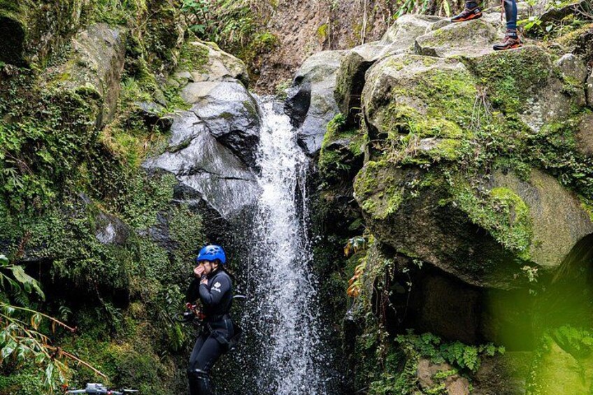 A person in a blue helmet and black clothing standing on a rock near a waterfall. 2