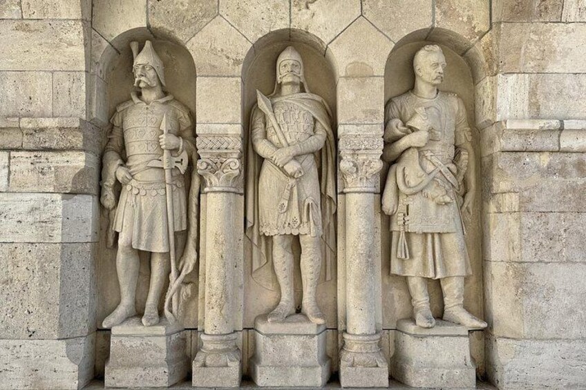 Three stone statues of armored figures in a row, each holding a weapon, set against a stone wall with arches. 3