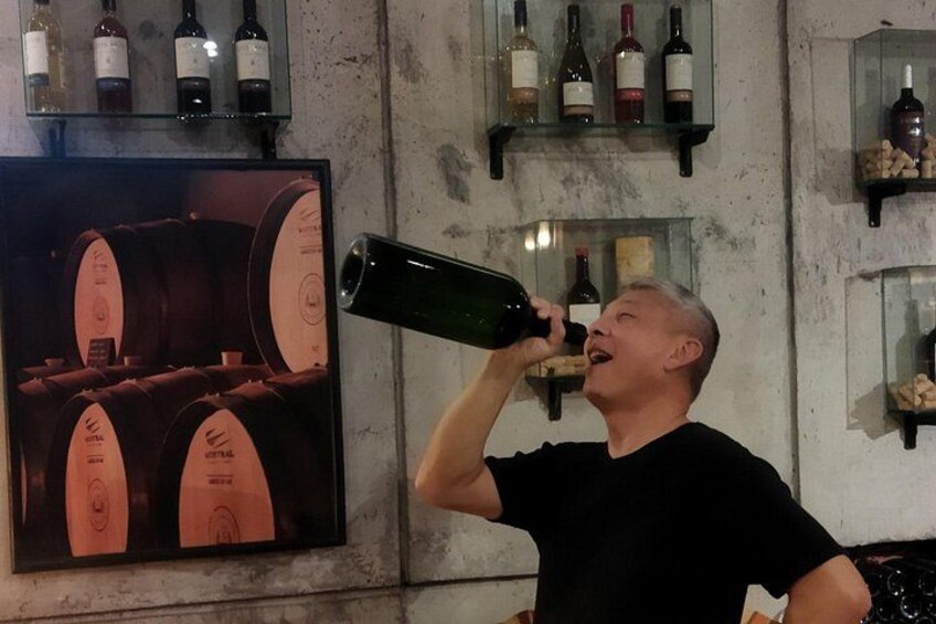 A person holding a wine bottle. There are shelves with wine bottles and a poster of wine barrels. 2
