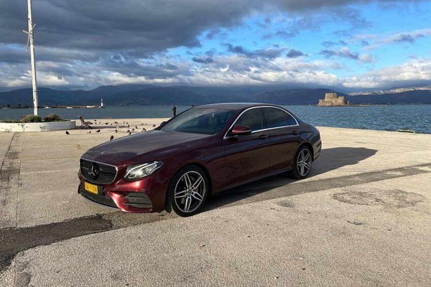 Un Mercedes-Benz color burdeos estacionado sobre una superficie de concreto cerca de una orilla marítima, con un cielo nublado y montañas al fondo. 4