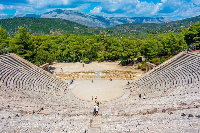 Un antiguo anfiteatro con asientos de piedra y un escenario central, rodeado de árboles y montañas al fondo. 5