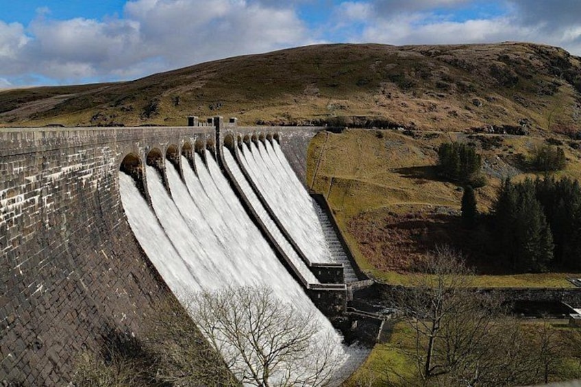 Een grote dam met meerdere overloopopeningen, omgeven door heuvelachtig terrein en schaarse begroeiing. 5