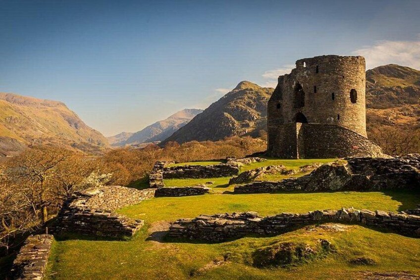 Een historisch stenen kasteel met ruïnes, omgeven door een grasrijk landschap en bergen op de achtergrond. 3