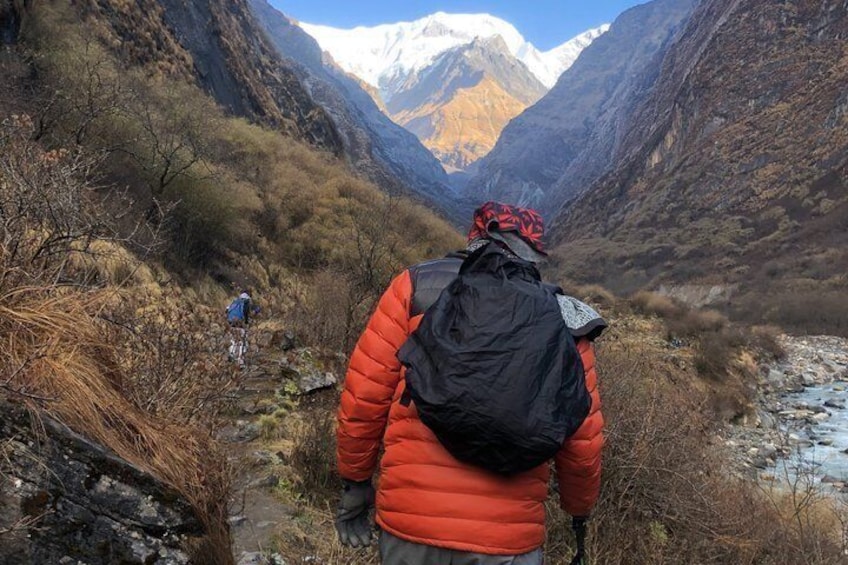 A hiker in a red jacket and black backpack walking on a rocky path in a mountainous area with a river and snow-capped peaks in the background. 2