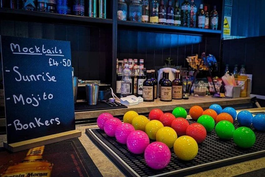 Un bar con un menú en pizarra que enumera cócteles sin alcohol y sus precios. Hay coloridas bolas en una bandeja frente al menú. 2