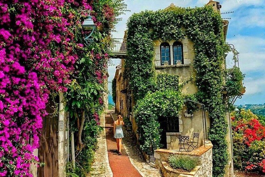 En smal gata med en byggnad täckt av grönska, en kvinna som går och bougainvilleablommor. 1