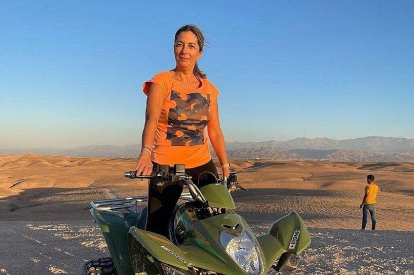 Une personne se tient à côté d’un quad vert dans un paysage désertique, avec des dunes de sable et des montagnes en arrière-plan. 3
