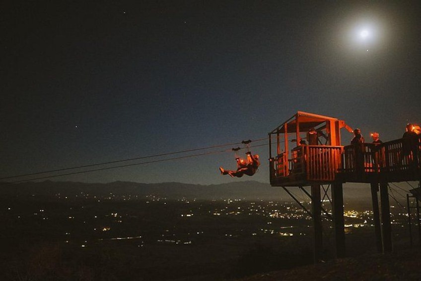 A night scene with a person swinging on a rope, a wooden platform, and a cityscape in the background. 1