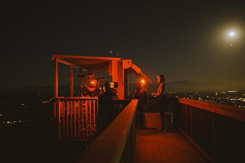 A nighttime scene with a wooden platform and a metal structure, possibly a stage or observation deck, overlooking a cityscape with lights. 4