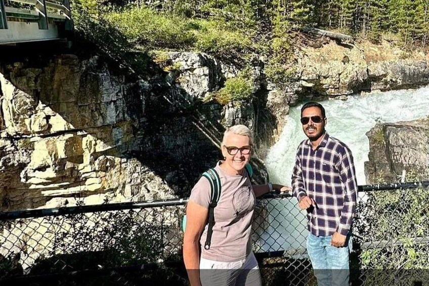Dos personas de pie junto a la orilla de un río rocoso, con una cascada al fondo. 5