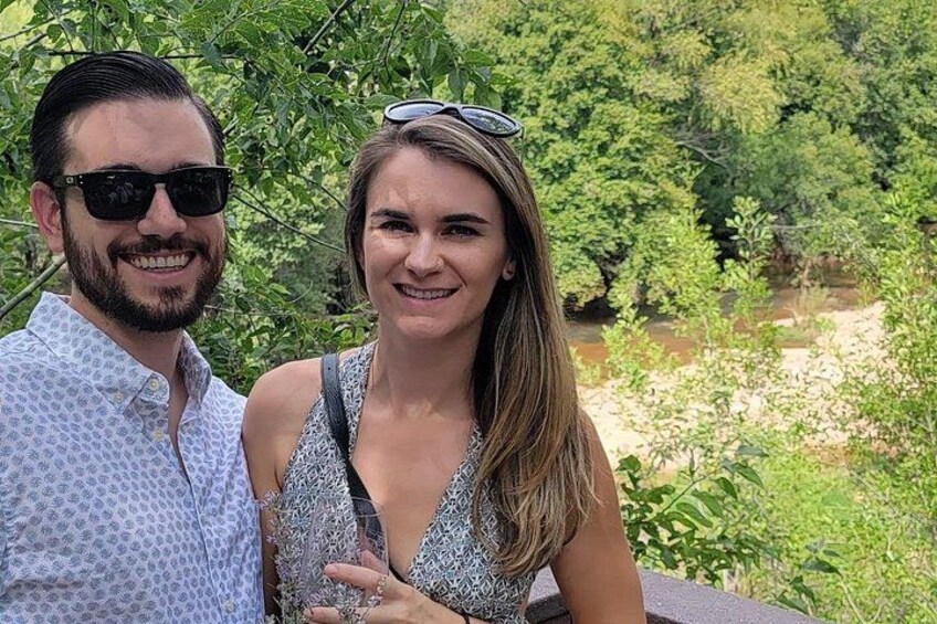 A person in a patterned dress and another in a light blue shirt standing outdoors with greenery and a river in the background. 3