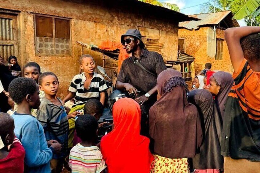 A group of people, including children, gathered around a man on a motorcycle in a rural setting with simple buildings in the background. 2