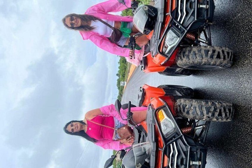 Two people on ATVs, one wearing a pink jacket, standing on a road with a cloudy sky in the background. 3