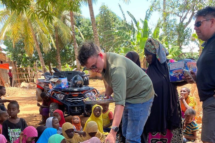 A man interacting with children near a vehicle, with palm trees and a rural setting in the background. 2