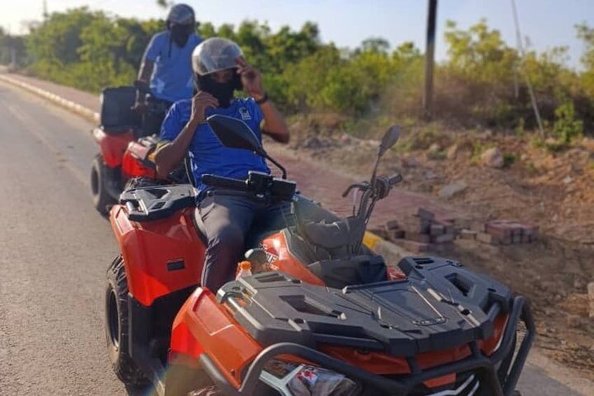 Two people on a red ATV on a paved road with vegetation in the background. 4