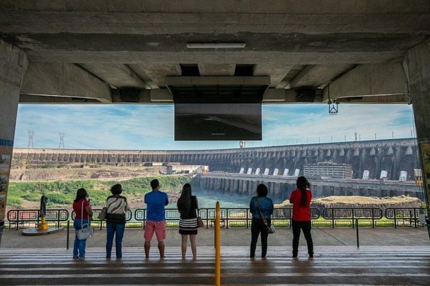 A group of people standing on a concrete platform, overlooking a large dam with multiple water gates. 5