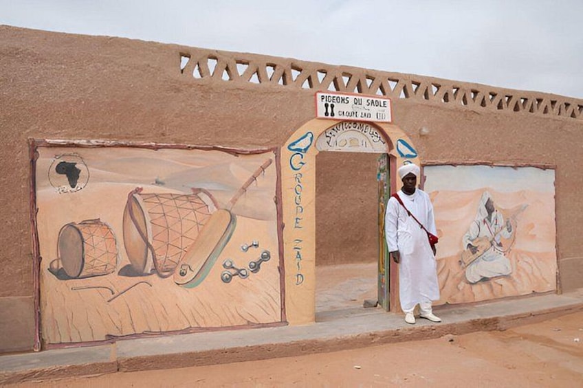 A man standing in front of a building with a sign reading 'Pigeons ou Sable' and a mural depicting drums and a figure. 3
