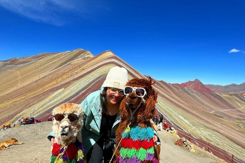 A person with a llama in a colorful, striped garment, standing in a mountainous area with a clear sky. 1