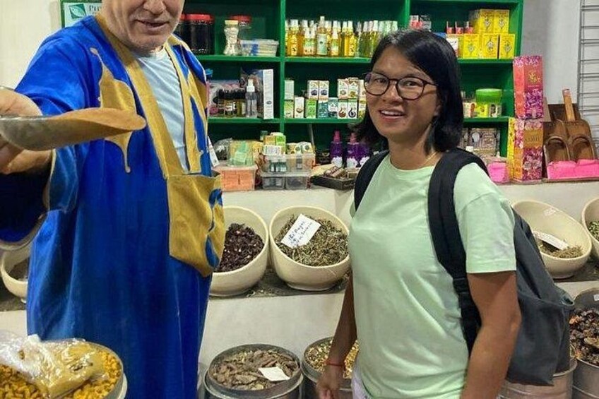 Eine Person in einem blauen Gewand mit gelbem Gürtel steht neben einer Person in einem grünen Hemd in einem Laden mit Regalen voller Produkte und großen Schalen mit Waren. 5
